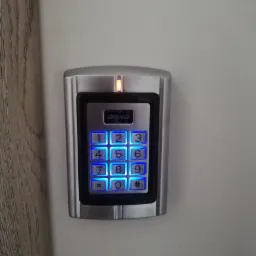 Klawiatura numeryczna domofonu ORNO z podświetlanymi na niebiesko przyciskami, zamontowana na szarej ścianie obok drewnianej futryny.