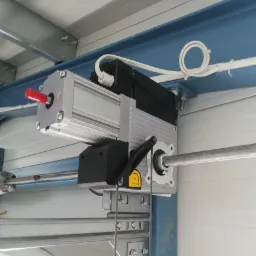 Zamontowany napęd elektryczny bramy garażowej z widocznym mechanizmem awaryjnego otwierania i kablem zasilającym, przymocowany do metalowej konstrukcji sufitu garażu.
