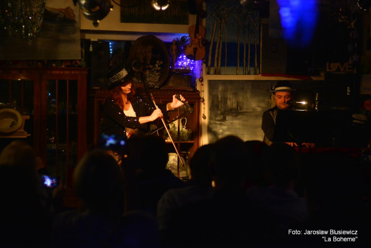 Kobieta w kapeluszu gra na wiolonczeli podczas występu na żywo, z pianistą w tle i publicznością obserwującą z przodu.