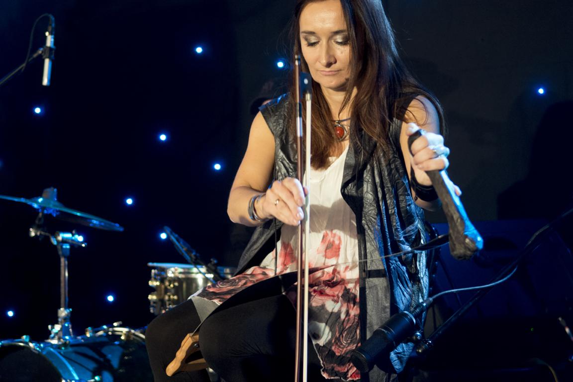 Kobieta gra na nietypowych skrzypcach elektrycznych na tle sceny z bębnami i gwiazdami.