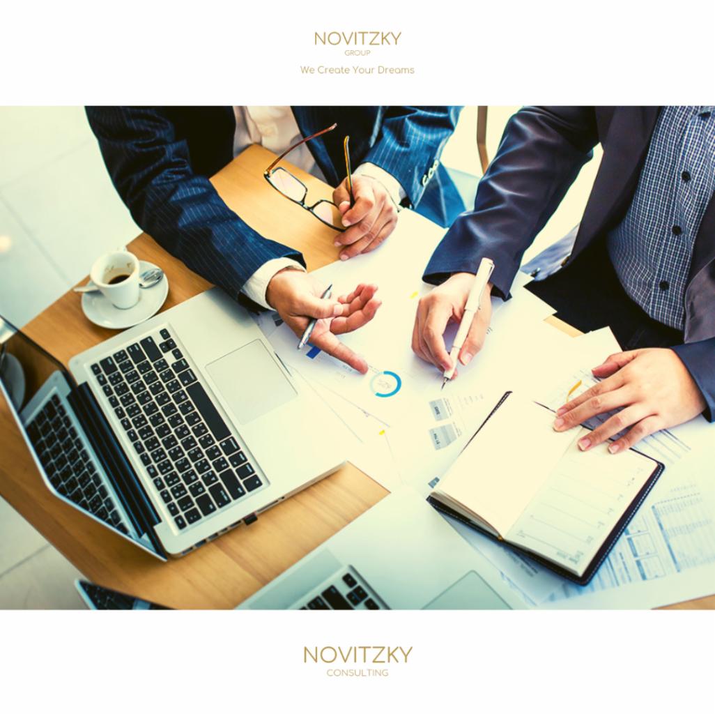 Dwóch mężczyzn w garniturach analizuje dokumenty i wykresy na stole z laptopami, notatnikiem i filiżanką kawy.