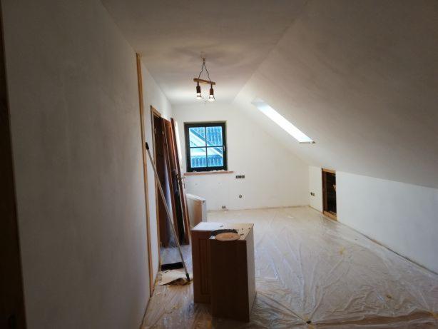 Poddasze w trakcie prac wykończeniowych: gładzie na ścianach i skośnym suficie, okno dachowe, zabezpieczona folią podłoga, drzwi w ościeżnicach o naturalnym odcieniu drewna oparte o ścianę...