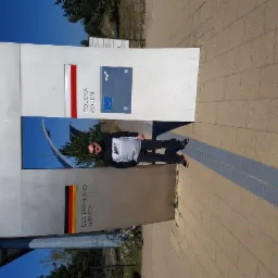 Mężczyzna w okularach i szarej koszulce z logo Nike stoi przy słupku granicznym z flagami Polski i Niemiec, na tle błękitnego nieba i kostki brukowej.