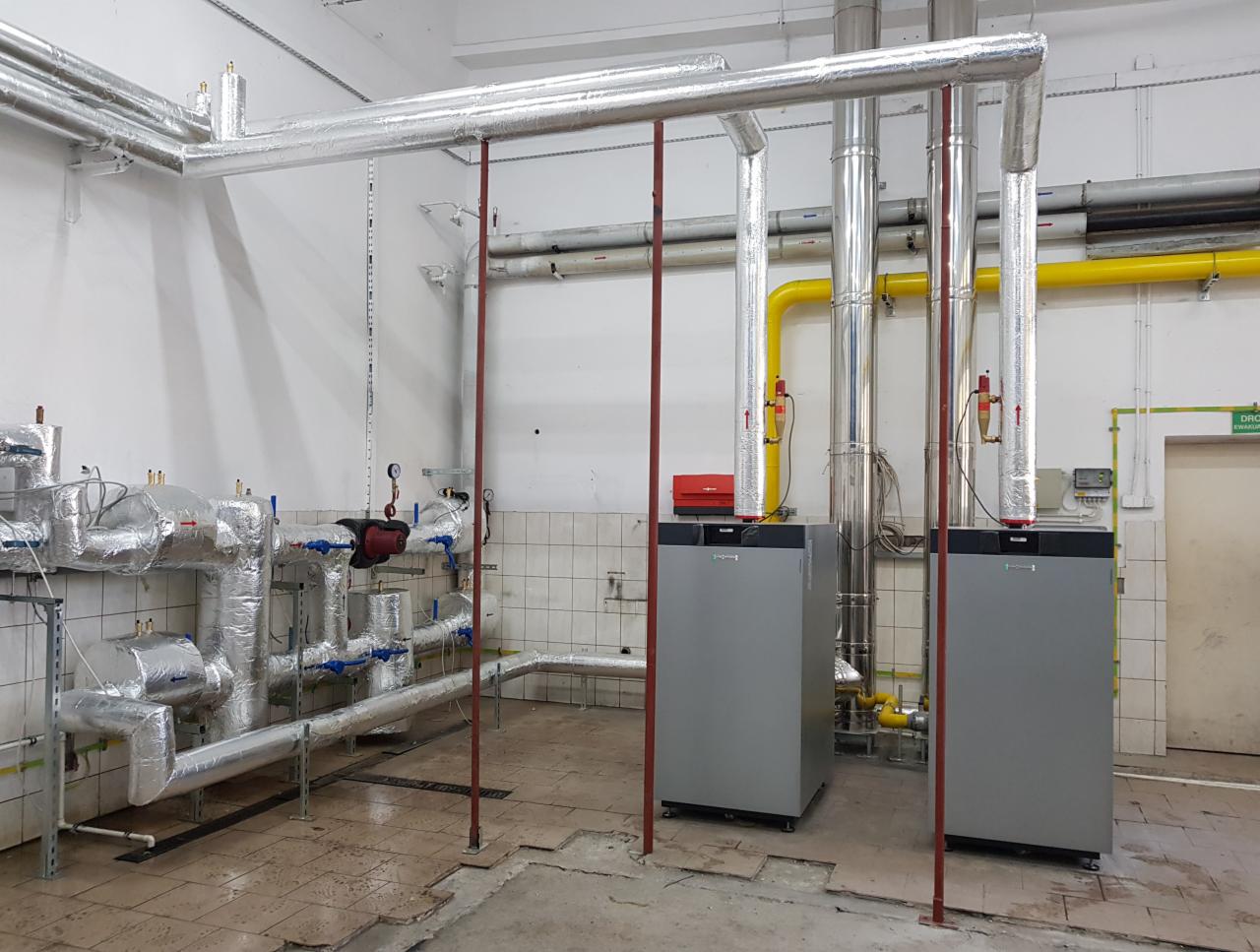 Dwa szare kotły grzewcze z rurami w izolacji aluminiowej, żółta rura gazowa, instalacja w pomieszczeniu z biało-szarą glazurą i betonową podłogą.