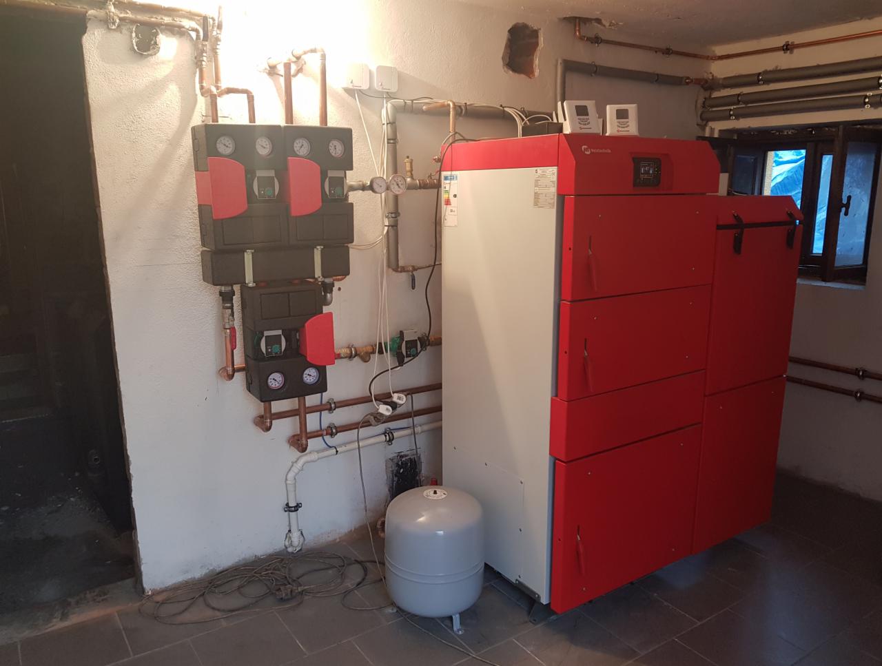 Zainstalowany, nowoczesny piec grzewczy z czerwonym frontem i szarą obudową, wraz z osprzętem i rurami miedzianymi na tle surowej ściany i szarej podłogi w piwnicy.