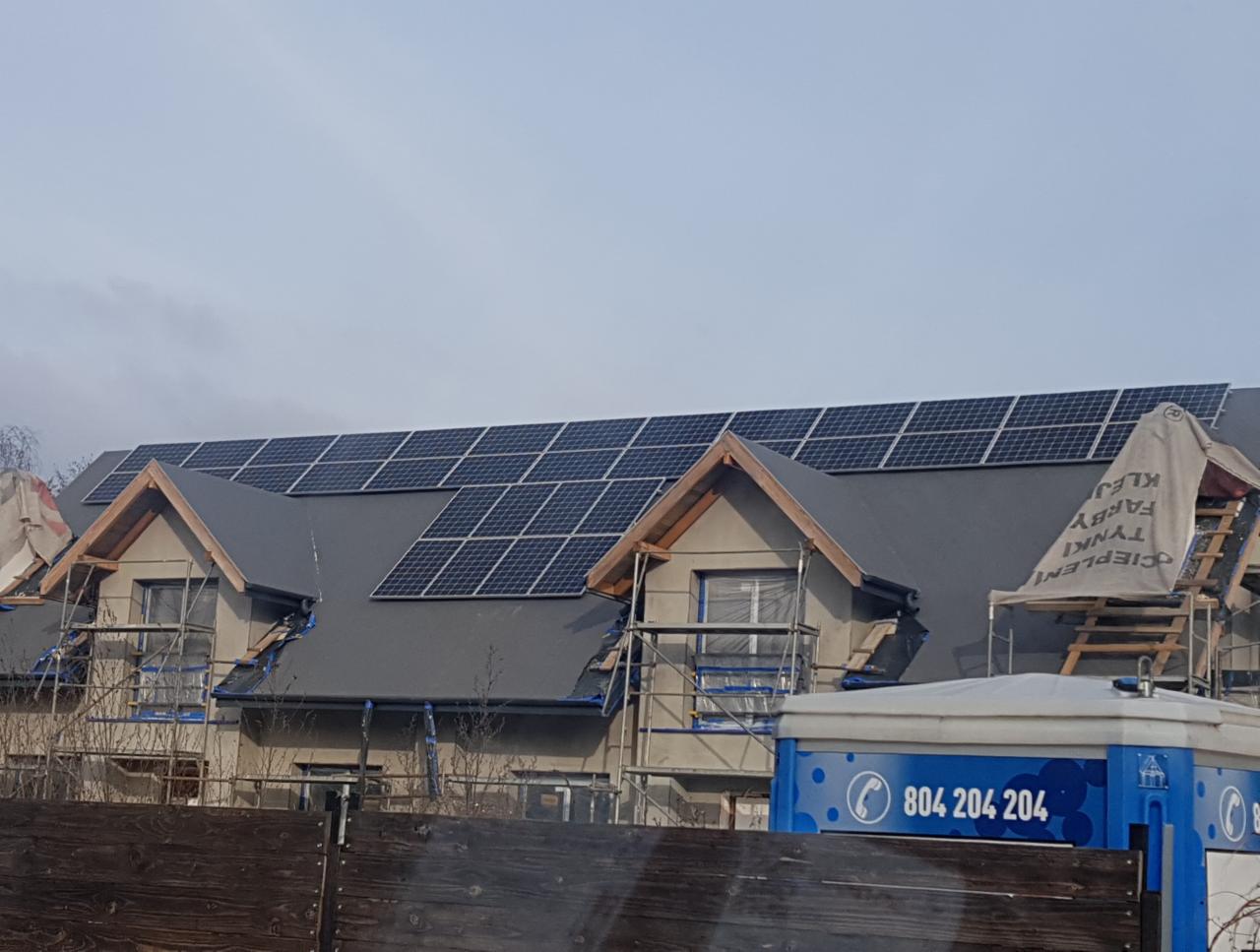 Montaż paneli słonecznych na dachu domu jednorodzinnego w trakcie budowy, widoczne rusztowania i zabezpieczenia okien folią, z dołu kadru widoczny fragment ogrodzenia i przenośna toaleta.