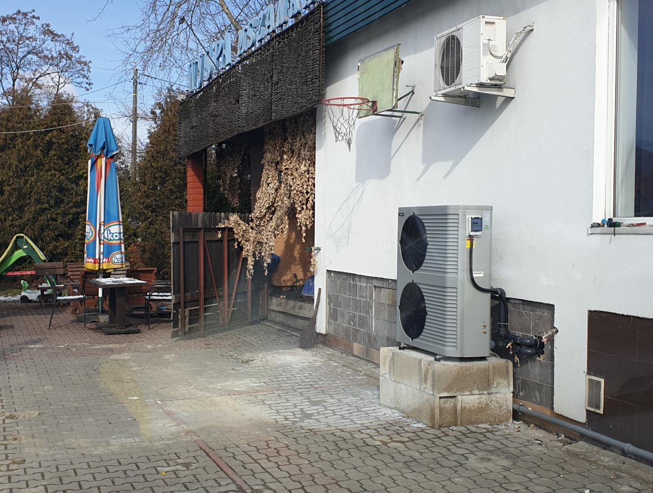 Zewnętrzna jednostka klimatyzacji dwuwentylatorowa na betonowym fundamencie przy ścianie budynku z zamontowaną jednostką wewnętrzną i koszem do koszykówki, widoczne rury instalacyjne.