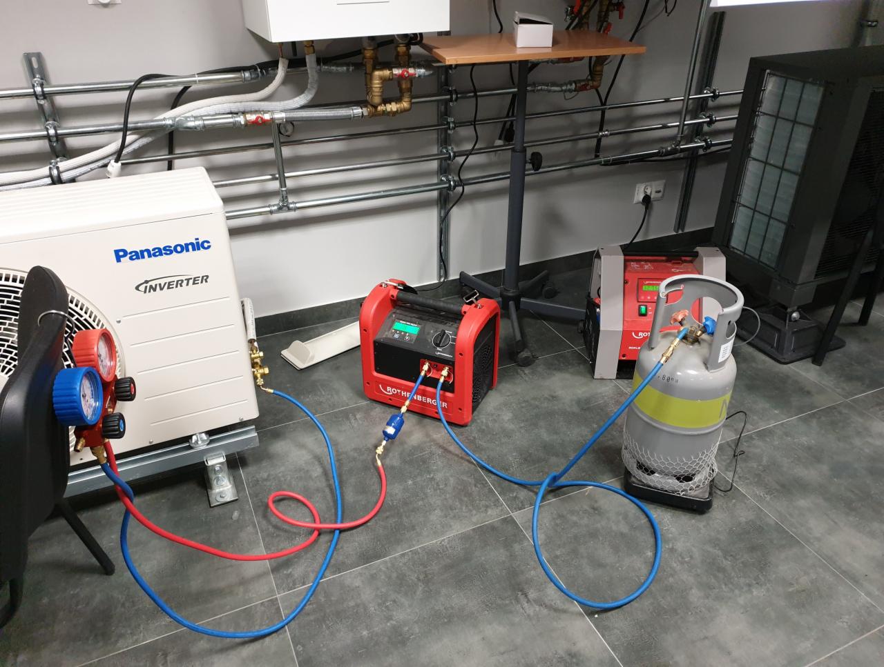 Sprzęt do serwisowania klimatyzacji: agregat Panasonic Inverter podłączony do butli z czynnikiem chłodniczym za pomocą manometrów i węży, obok czerwona stacja do odzysku czynnika.