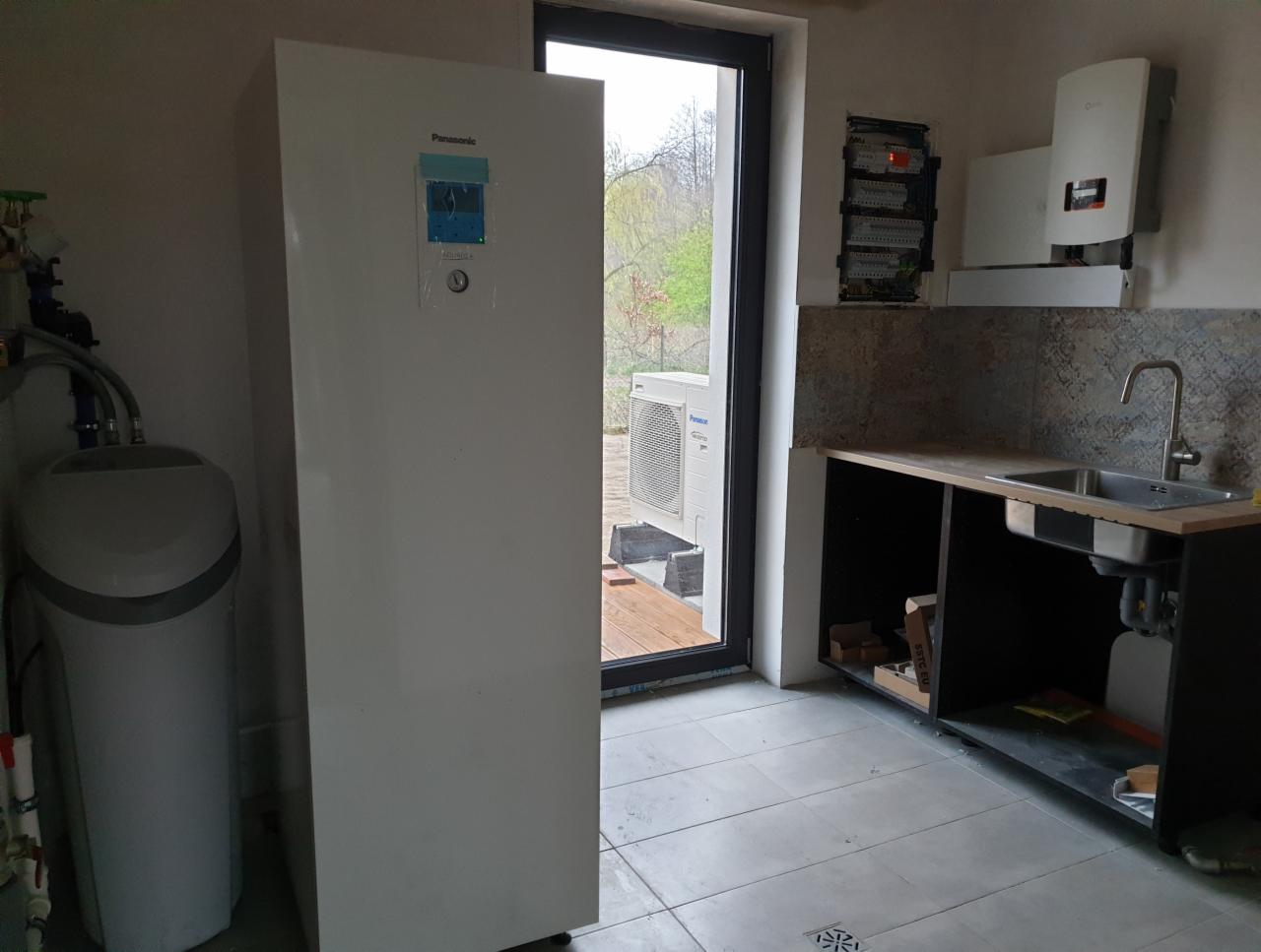 Wnętrze pomieszczenia z pompą ciepła Panasonic, zmiękczaczem wody, instalacją elektryczną i zlewem, widok przez okno na zewnątrz.