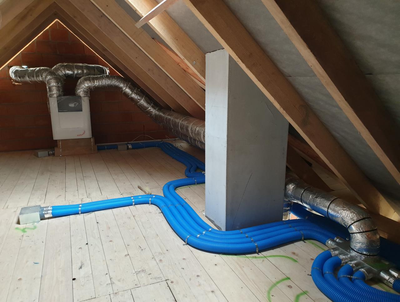 Instalacja rekuperacji na poddaszu: centrala wentylacyjna z izolowanymi przewodami wentylacyjnymi w srebrnej folii oraz niebieskie rury rozprowadzające powietrze, widoczne elementy konstrukcji dachu.