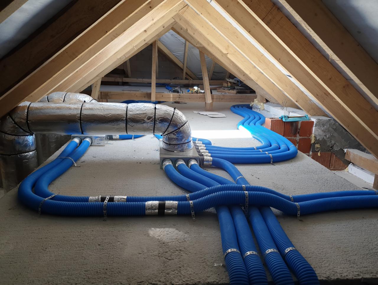 Niebieskie, karbowane rury wentylacyjne rozprowadzone na poddaszu, pod drewnianą konstrukcją dachu, z izolowanymi przewodami i skrzynką rozdzielczą.