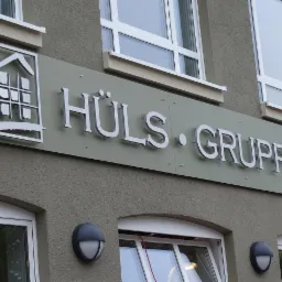 Zewnętrzny szyld firmy HÜLS GRUPPE z logo przedstawiającym schematyczny dom z oknami, zamontowany na szarej elewacji budynku.