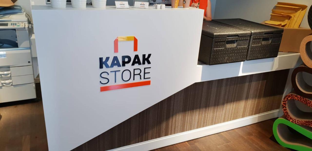 Białe biurko recepcyjne z logo 'Kapak Store' w kolorach czerwonym, żółtym i czarnym. Za biurkiem widoczne pojemniki termoizolacyjne i inne akcesoria.