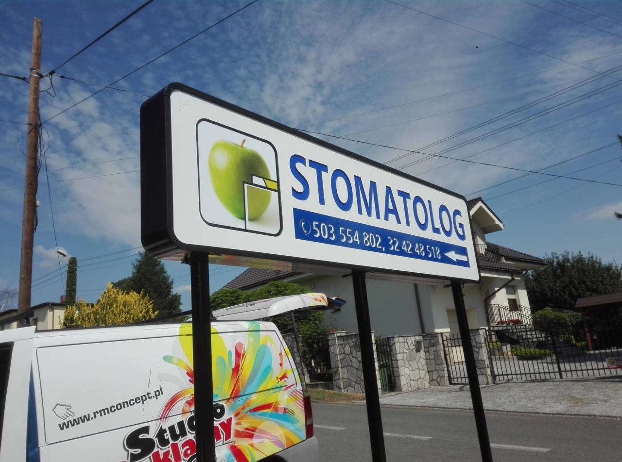 Szyld reklamowy gabinetu stomatologicznego z grafiką zielonego jabłka i numerami telefonów, widoczny z ulicy, z fragmentem firmowego samochodu w tle.