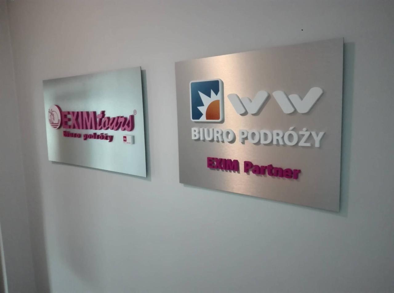 Dwie metalowe tablice informacyjne wiszące na ścianie: jedna z logo 'EXIMtours' i hasłem 'biuro podróży', druga z logo i napisem 'BIURO PODRÓŻY EXIM Partner'.