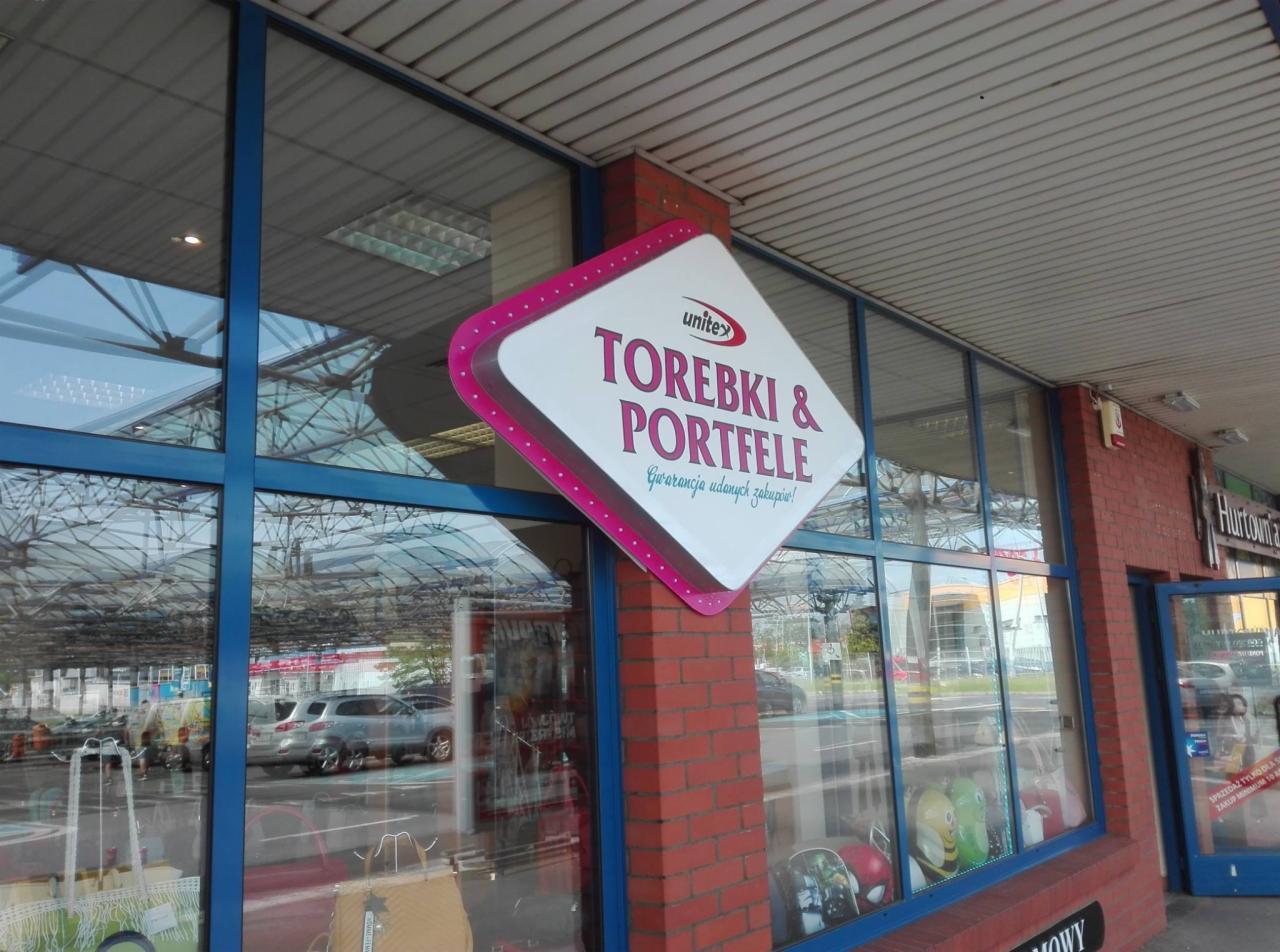 Zewnętrzny szyld sklepu z torebkami i portfelami, umieszczony na ceglanym filarze przy wejściu, z widoczną witryną sklepową i zaparkowanymi samochodami w tle.