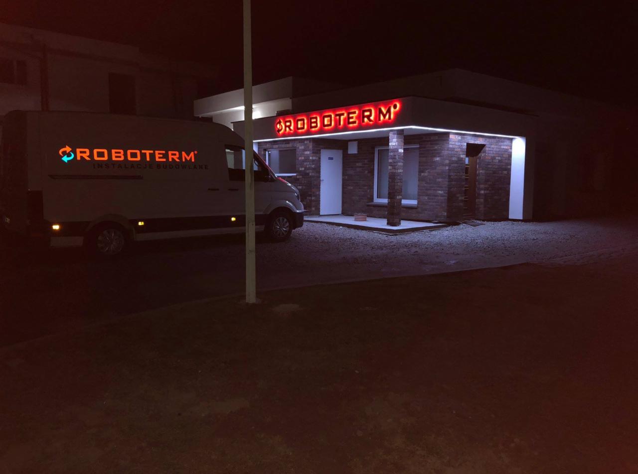 Nocny widok na budynek firmy z podświetlonym neonowym logo na fasadzie oraz samochód dostawczy z logo firmy, oświetlony z boku.