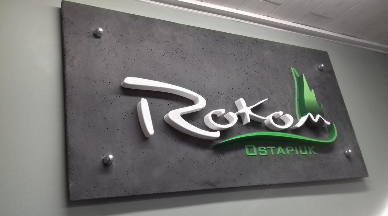 Tablica firmowa z betonu architektonicznego z trójwymiarowym białym napisem i zielonym logo, zamontowana na ścianie za pomocą metalowych dystansów.