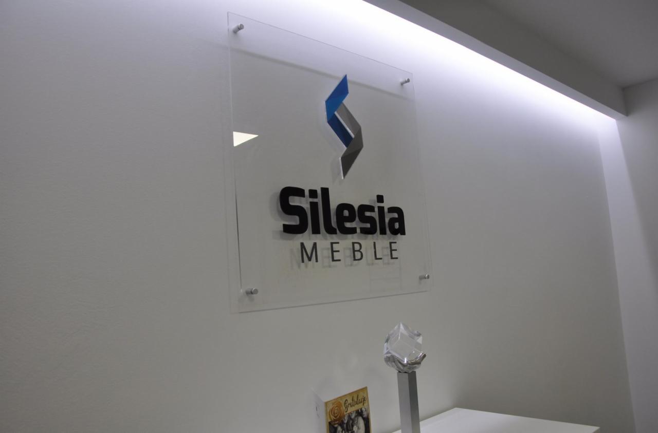 Szklana tablica z logo i nazwą firmy Silesia Meble, zamocowana na ścianie, podświetlona od góry.