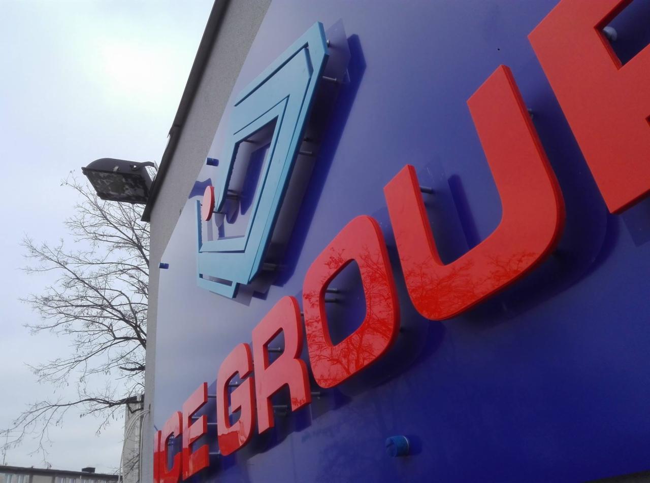 Zewnętrzny szyld z logo firmy w kolorach czerwonym i niebieskim, zamontowany na niebieskiej płycie, widoczny z dołu pod kątem, z odbiciem drzew na literach.