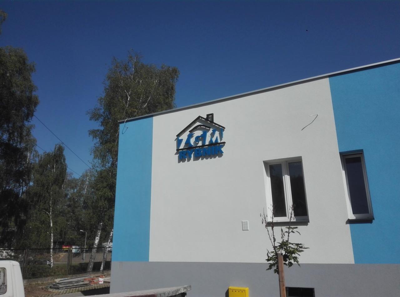Elewacja budynku z niebieskimi pasami i zamontowanym niebieskim logo firmy 'ZGM Rybnik' nad oknem, widok z dołu.