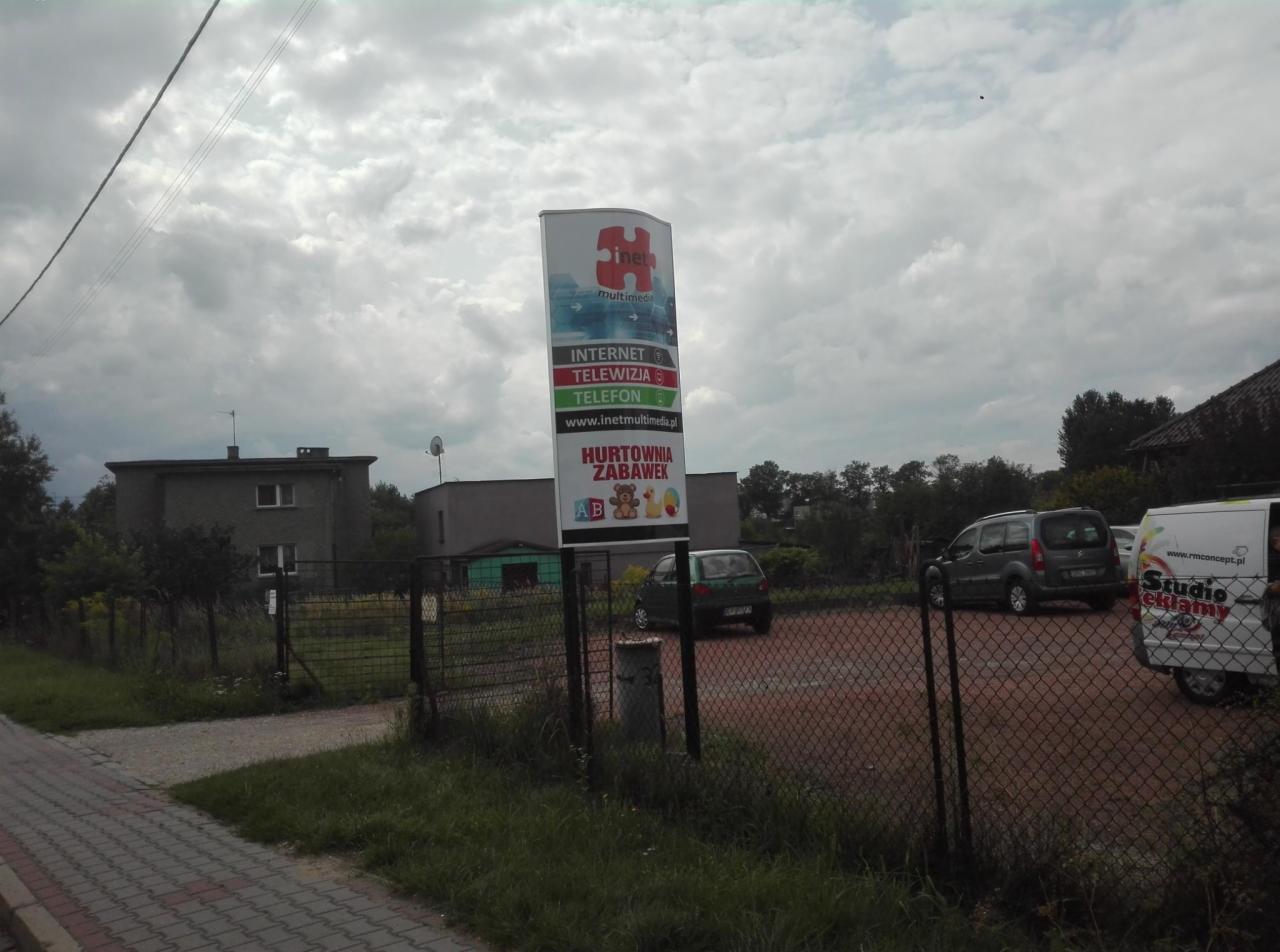 Pionowy szyld reklamowy firmy telekomunikacyjnej z logo, adresem strony internetowej i informacją o hurtowni zabawek, umieszczony na parkingu przed budynkiem.