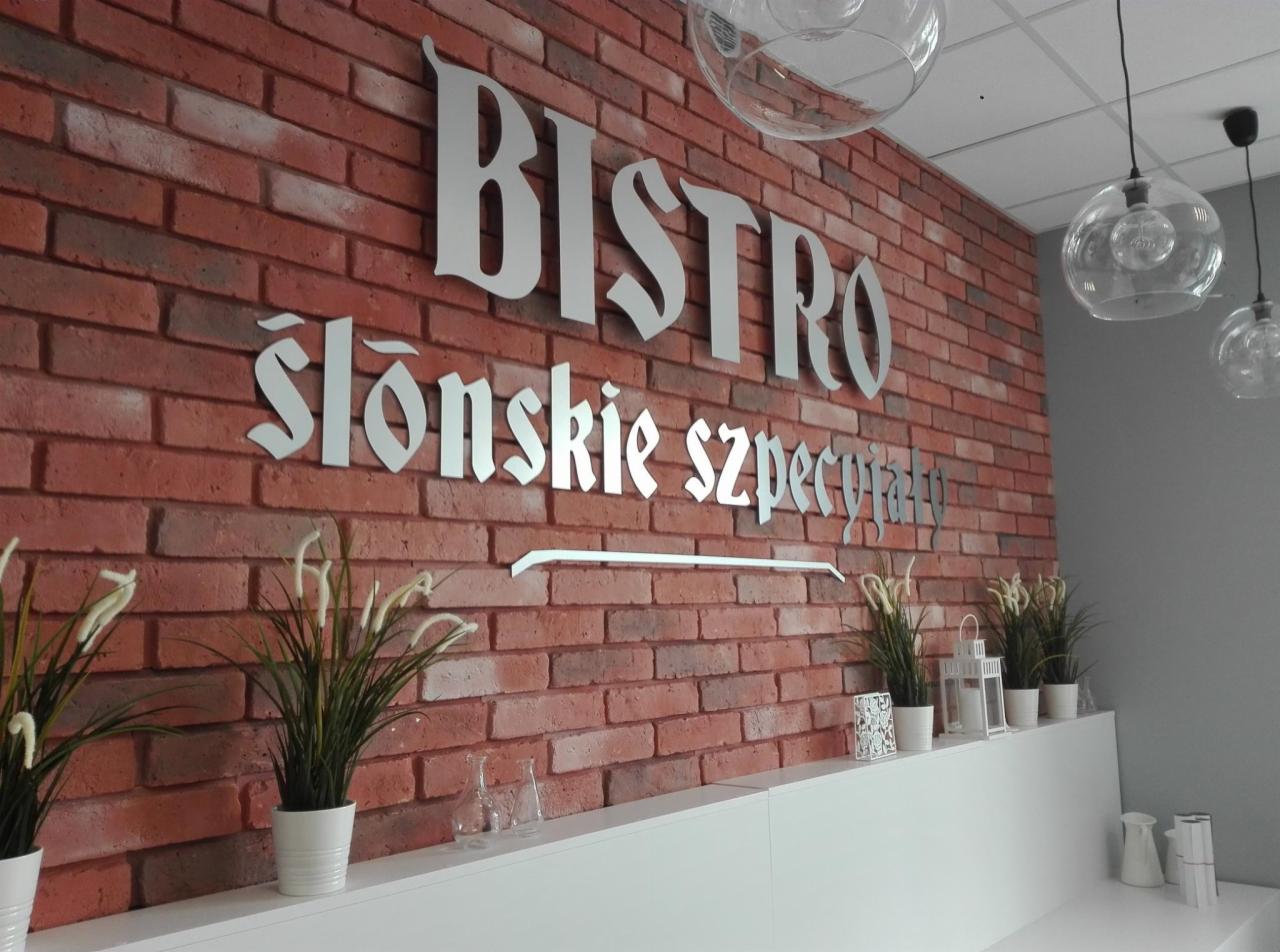Metalowe litery tworzące napis 'Bistro Ślonskie Specjały' na ścianie z czerwonej cegły, dekoracyjne rośliny w białych doniczkach na białej półce, szklane lampy wiszące.
