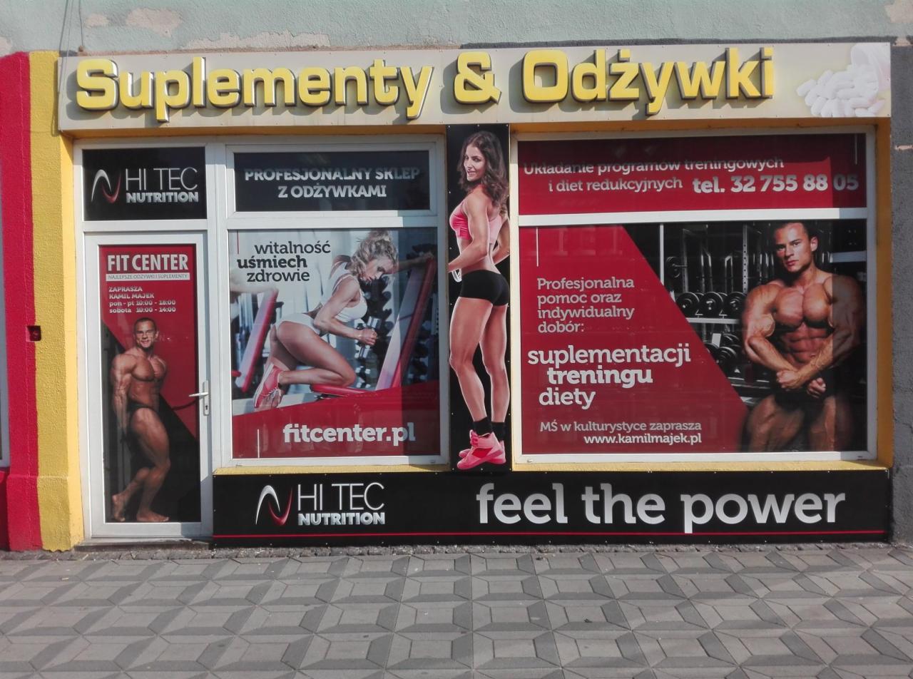 Fasada sklepu z suplementami i odżywkami, z reklamami w oknach prezentującymi sylwetki sportowców i hasła związane z treningiem i dietą.