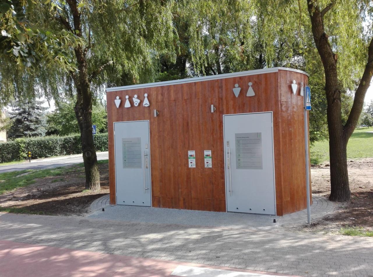 Nowoczesny, drewniany pawilon sanitarny z dwoma wejściami, oznaczonymi piktogramami płci, umieszczony w przestrzeni parkowej z drzewami i ścieżką rowerową.