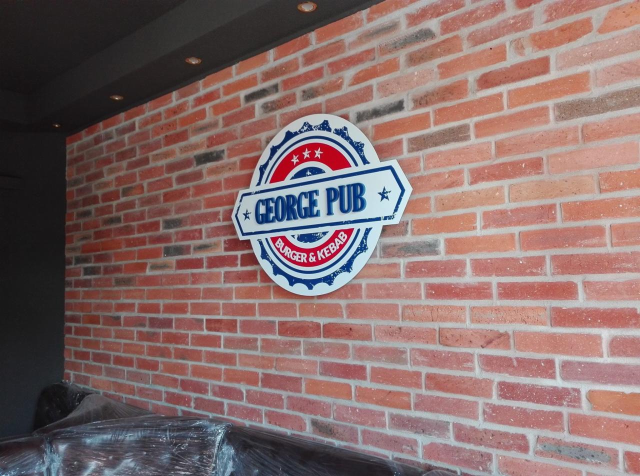 Szyld reklamowy 'George Pub Burger & Kebab' na ceglanej ścianie, z elementami w kolorach czerwonym, białym i niebieskim, w stylu retro.