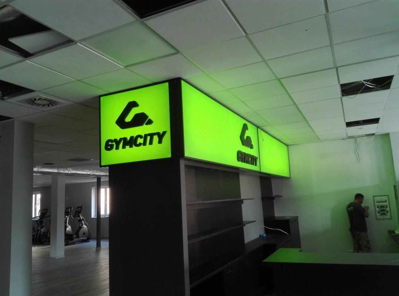 Podświetlane kasetony z logo 'Gymcity' zamontowane pod sufitem w lokalu, widoczne elementy wyposażenia siłowni w tle.