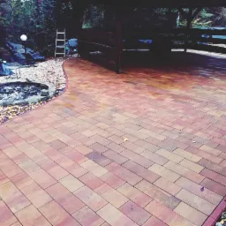 Nowo ułożony, wzorzysty chodnik z kostki brukowej w odcieniach czerwieni i brązu, prowadzący do drewnianej altany i okrągłego paleniska otoczonego kamieniami. Widoczne narzędzia pracy w skrzynce.