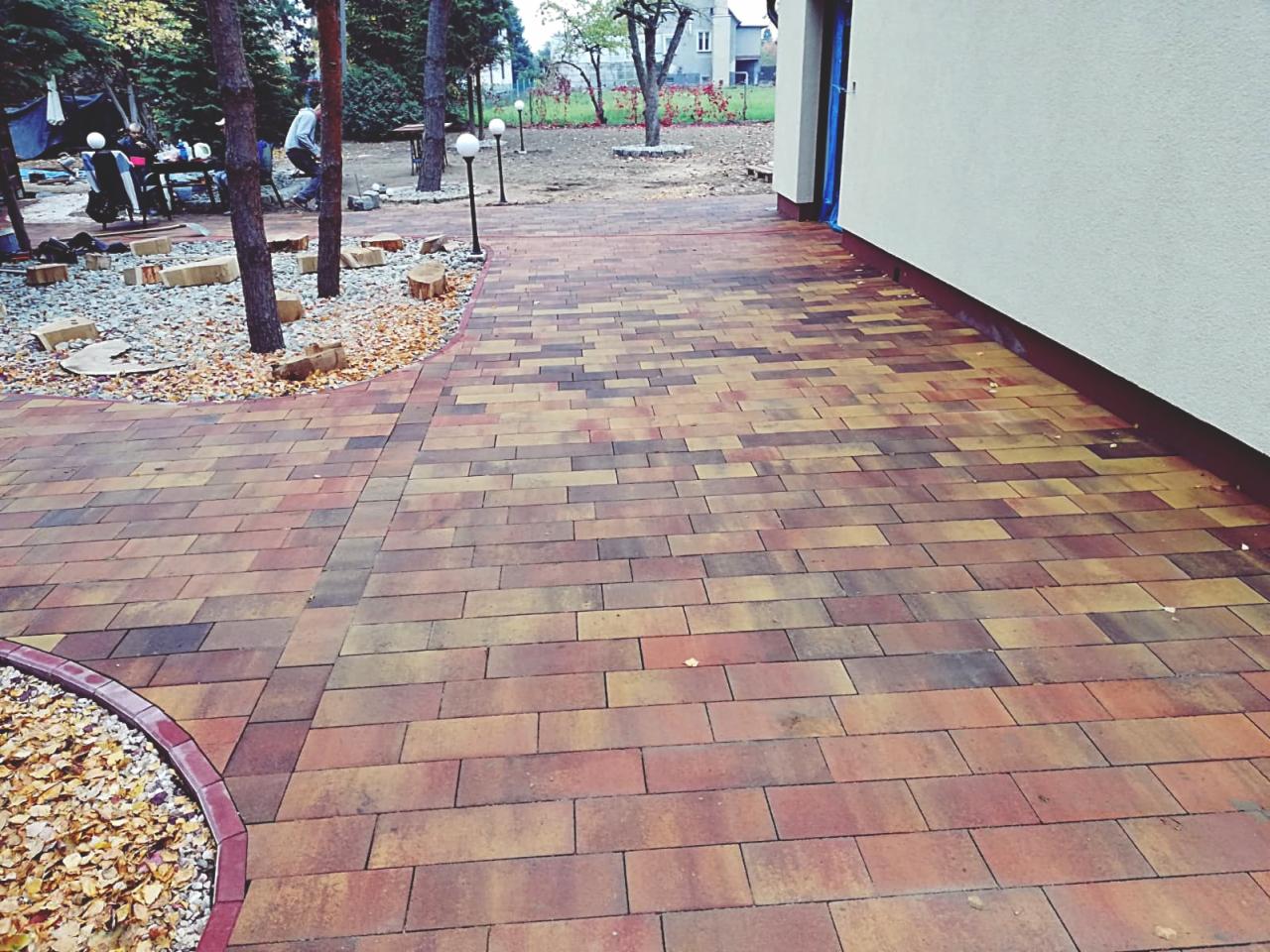 Ułożona kostka brukowa w odcieniach brązu i żółci, tworząca ścieżkę w ogrodzie, z widocznym obrzeżem z kostki i fragmentem trawnika z drzewami w tle.