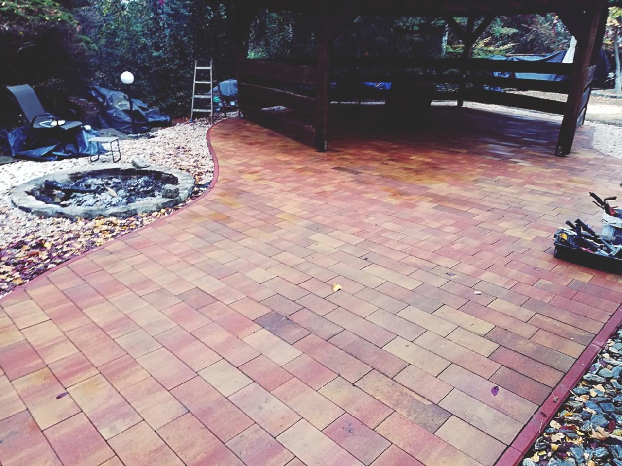 Nowo ułożony, wzorzysty chodnik z kostki brukowej w odcieniach czerwieni i brązu, prowadzący do drewnianej altany i okrągłego paleniska otoczonego kamieniami. Widoczne narzędzia pracy w skrzynce.
