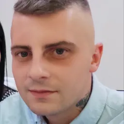 Portret mężczyzny z krótkimi włosami i tatuażem na szyi, ubrany w jasnoniebieską koszulę, na jasnym tle.