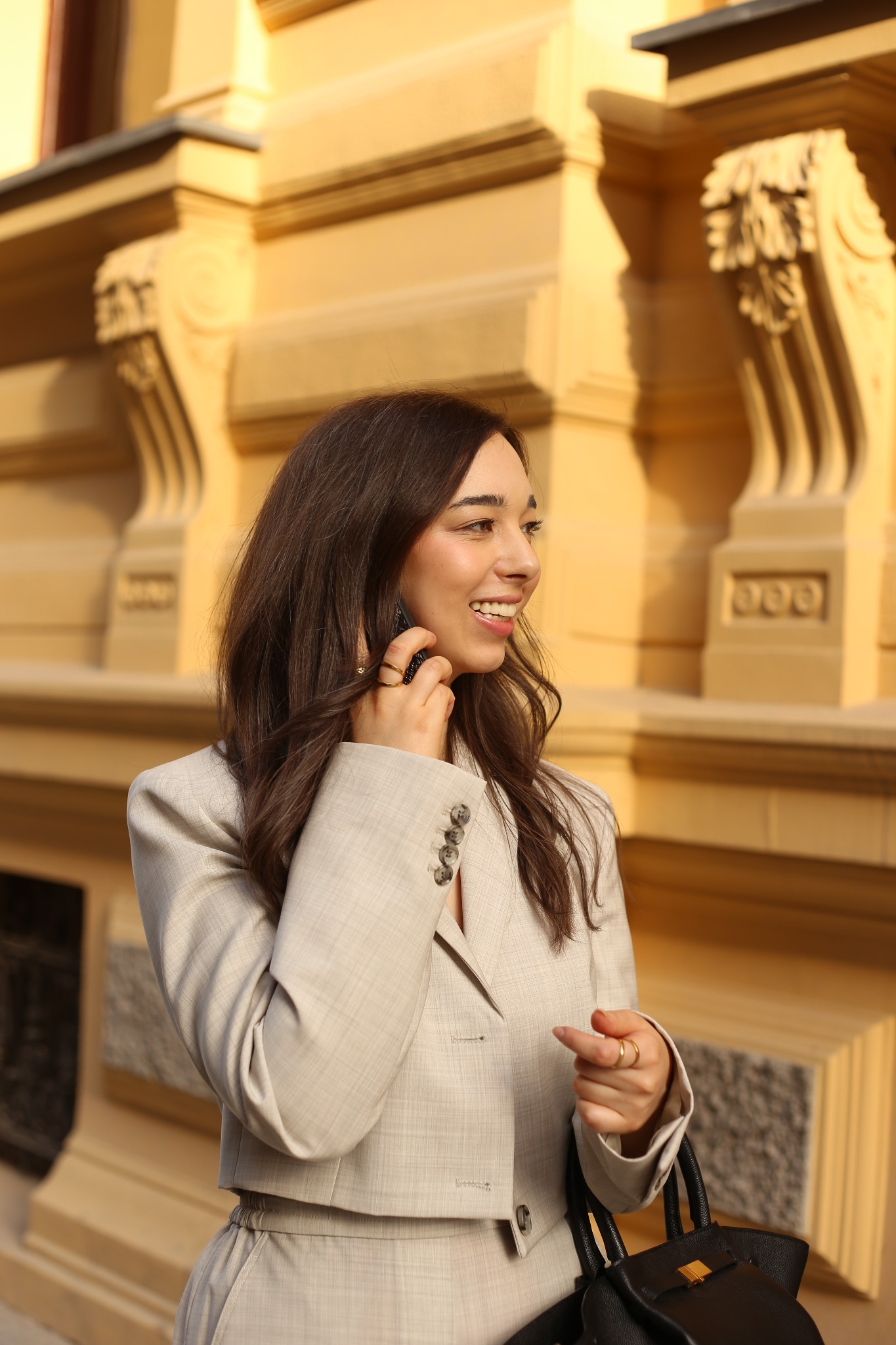 Uśmiechnięta brunetka w beżowym, dopasowanym garniturze rozmawia przez telefon na tle architektonicznej fasady budynku.