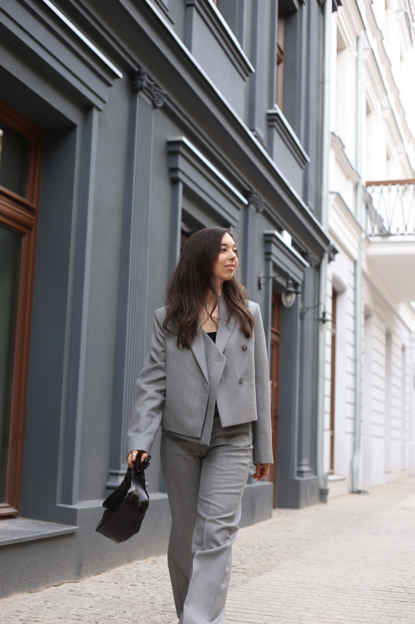 Kobieta w szarym, dwurzędowym garniturze spaceruje ulicą z torebką w dłoni, budynki w tle z szarymi i białymi fasadami.