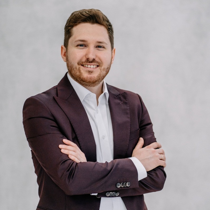 Mężczyzna w bordowym garniturze i białej koszuli, z brodą, uśmiechnięty, pozujący z założonymi rękami na jasnym tle.