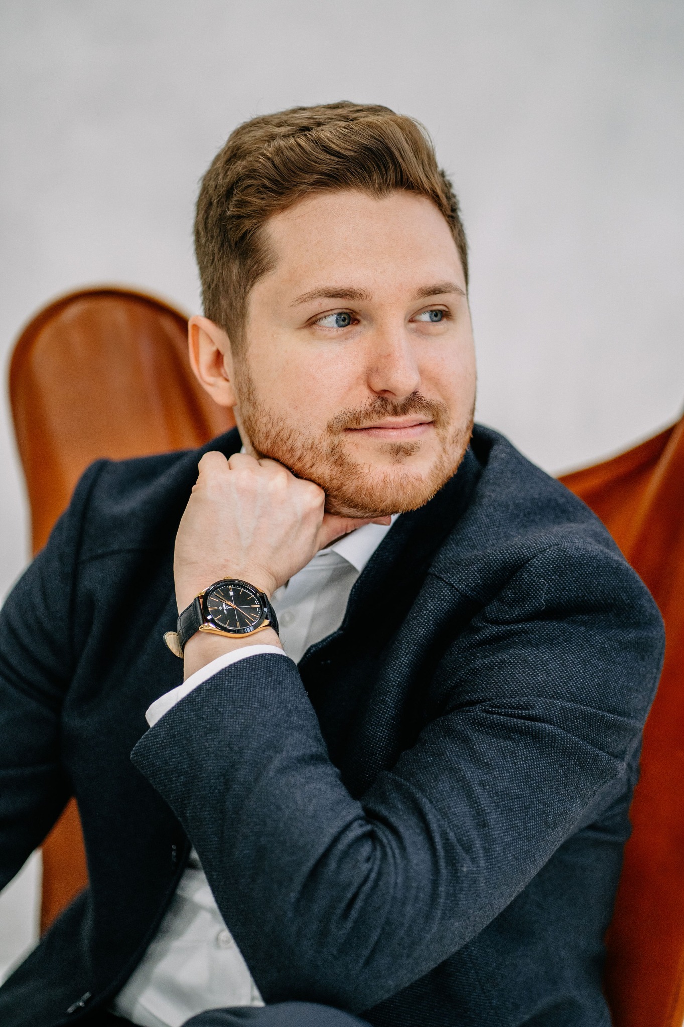 Portret mężczyzny w garniturze, z rudą brodą, opierającego podbródek na dłoni z zegarkiem na skórzanym pasku, siedzącego w skórzanym fotelu.