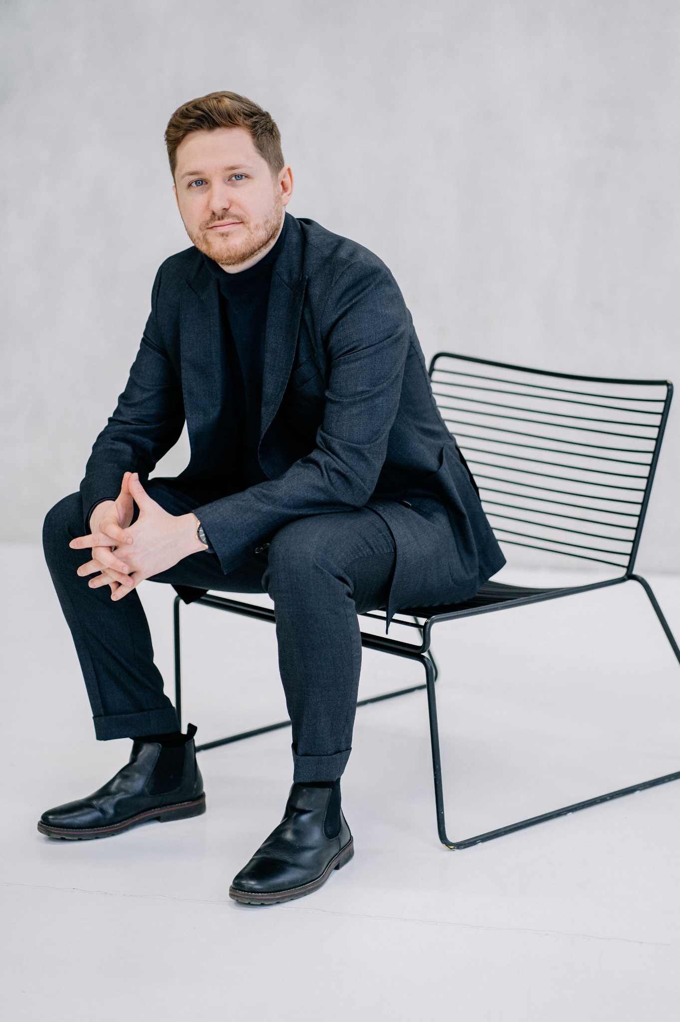 Portret mężczyzny w ciemnym garniturze i czarnym golfie, siedzącego na metalowym krześle na białym tle, z dłońmi splecionymi na kolanach i widocznym zegarkiem na nadgarstku.