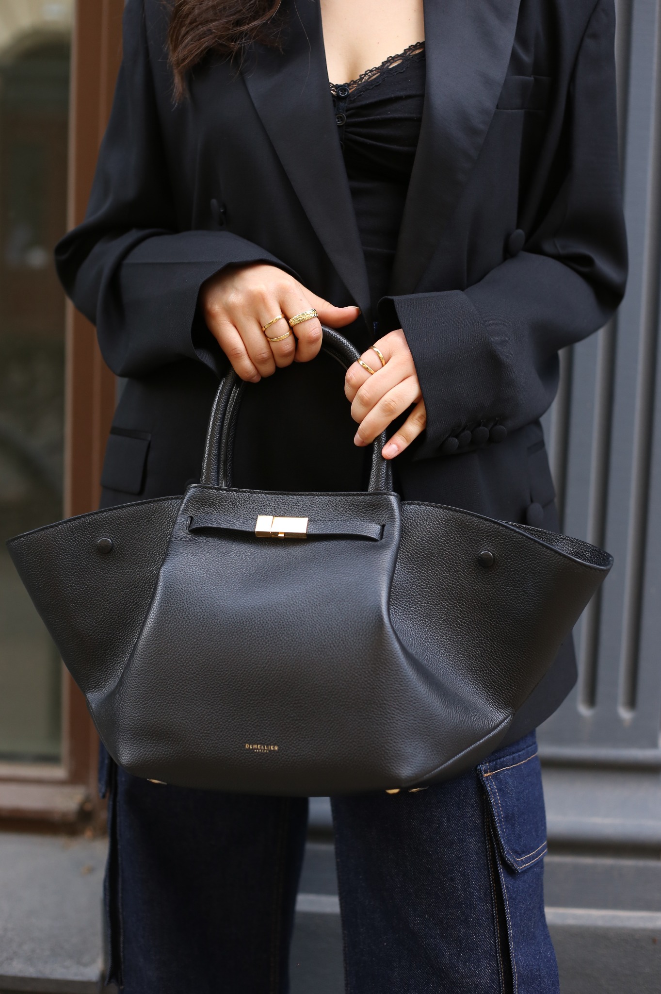 Kobieta w czarnej marynarce trzyma czarną skórzaną torebkę z złotymi detalami i pierścionkami na palcach.