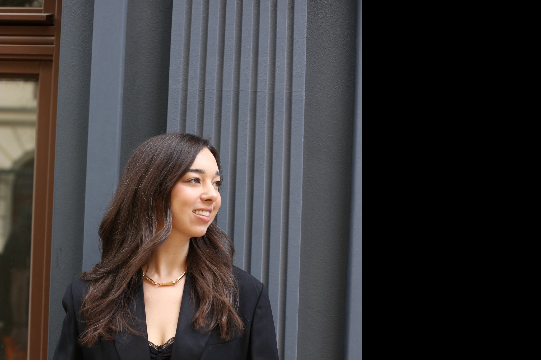 Uśmiechnięta kobieta z brązowymi, falowanymi włosami, ubrana w czarną marynarkę i złotą kolie, pozuje na tle szarej, pionowo żłobkowanej ściany.