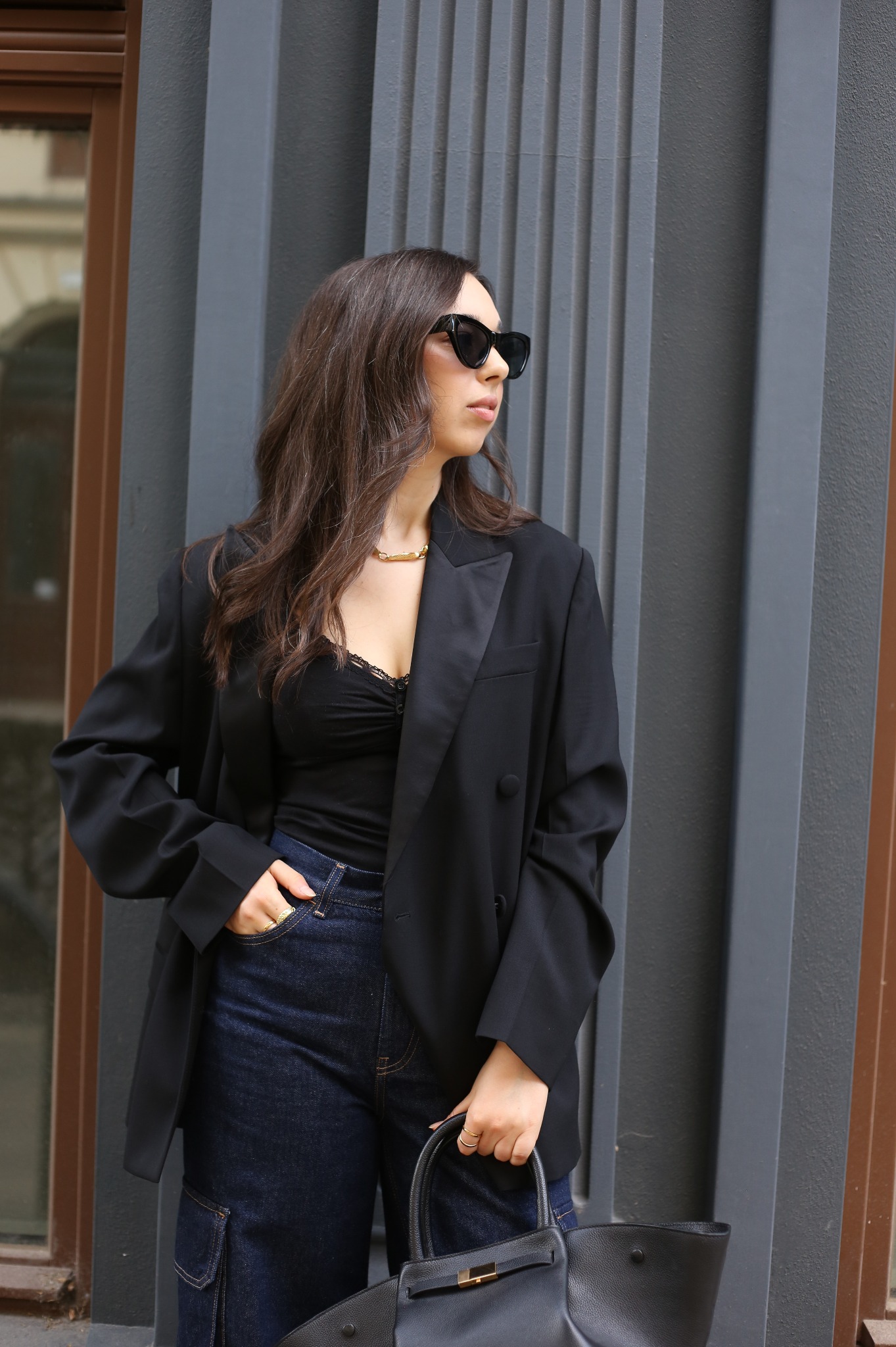 Kobieta w czarnej marynarce, topie z koronką i ciemnych jeansach cargo, z czarną torebką i okularami przeciwsłonecznymi, pozująca na tle szarej ściany z pionowymi żłobieniami.