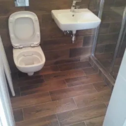 Wykończona łazienka z brązowymi płytkami imitującymi drewno na podłodze i ścianach, biała umywalka narożna i toaleta z otwartą klapą, kabina prysznicowa w rogu.