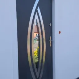 Nowoczesne, grafitowe drzwi wejściowe z dekoracyjnymi, srebrnymi wstawkami w kształcie łzy, zamontowane w białej ścianie budynku.