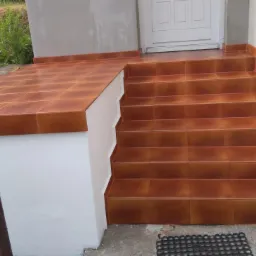 Brązowe płytki ceramiczne na schodach wejściowych i podeście, białe ściany, widoczne elementy drewnianego płotu w tle.