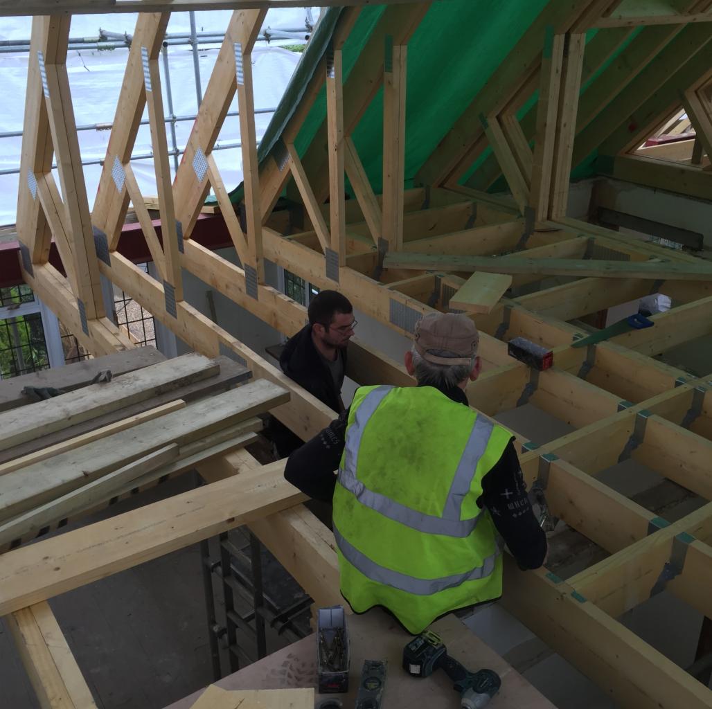 Dwóch pracowników budowlanych montuje drewnianą konstrukcję więźby dachowej, widoczne elementy metalowe łączące belki, jeden pracownik w żółtej kamizelce odblaskowej, drugi w ciemnej bluzie, w tle...