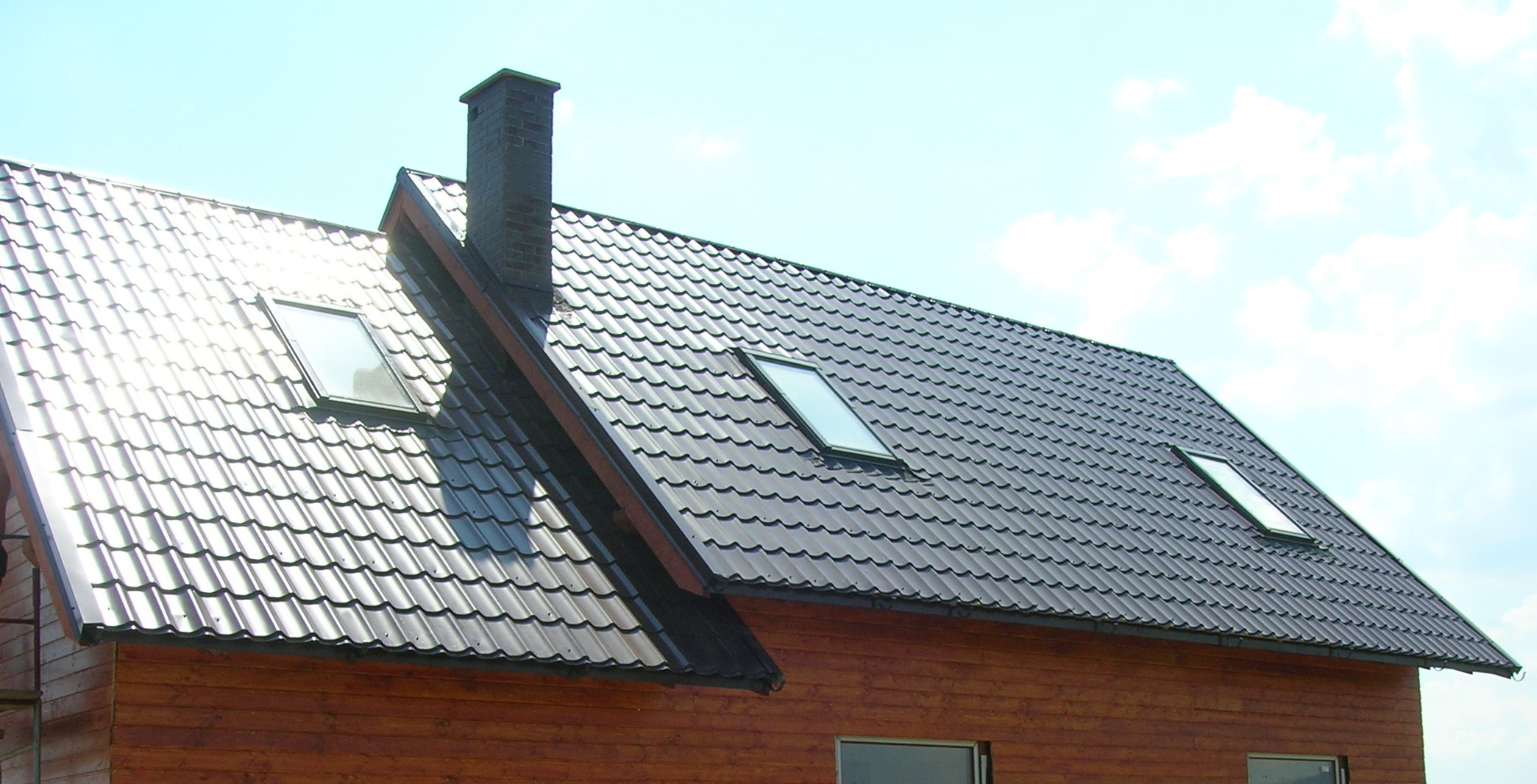 Kompletny dach z brązowej blachodachówki, z trzema oknami dachowymi i kominem z cegły, na tle błękitnego nieba.