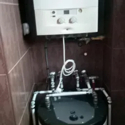 Biały kocioł gazowy wiszący nad cylindrycznym zasobnikiem wody z podłączeniami hydraulicznymi w pomieszczeniu wyłożonym brązowymi płytkami.