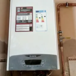 Biały, wiszący kocioł gazowy Ariston ONE z podłączonym kominem i miedzianymi rurami na tle beżowej ściany i kafelkowanej półki.